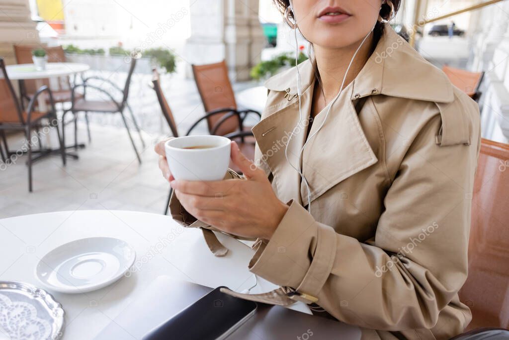 cropped view of woman in trench coat listening music and holding cup near smartphone and laptop in cafe terrace