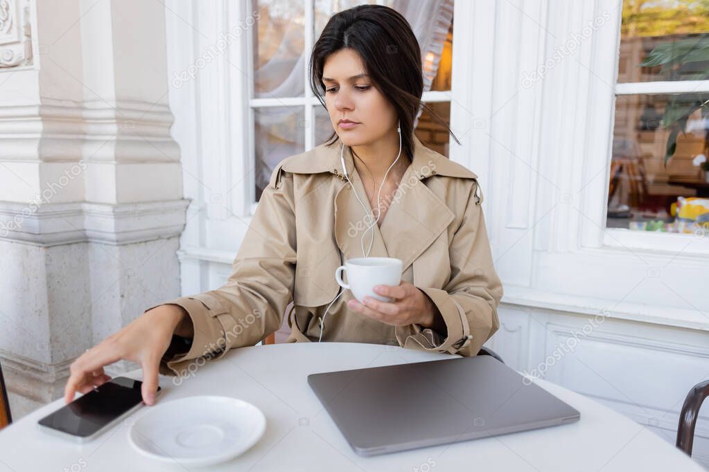 brunette woman in trench coat listening music and holding cup while reaching smartphone on table in cafe terrace