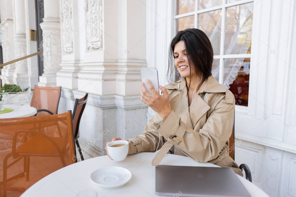smiling woman in trench coat listening music and holding cup while taking photo on smartphone in cafe terrace