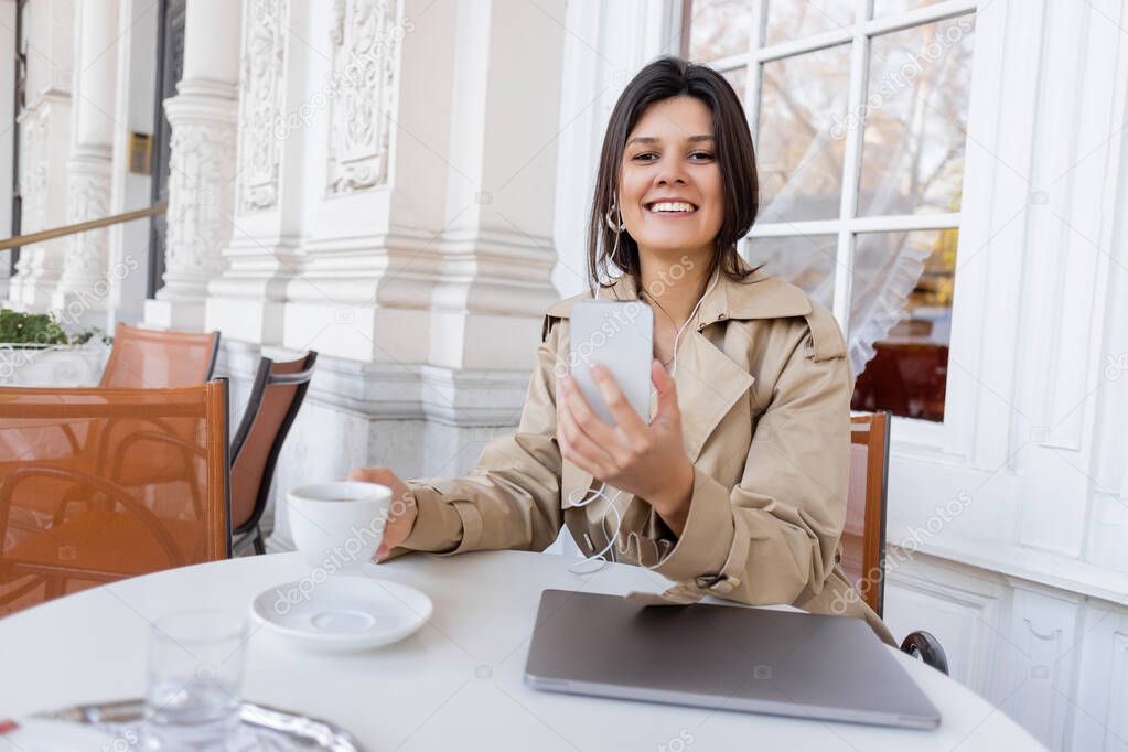 smiling woman in trench coat listening music in earphones and holding cup while using smartphone in cafe terrace