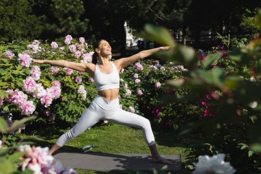 Savaşçı pozu veren mutlu, sportif bir kadın. Parkta uzanmış elleri var. 