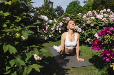 Yeşil çiçek açan bitkilerin yanındaki yoga minderinde poz veren pozitif kadın.