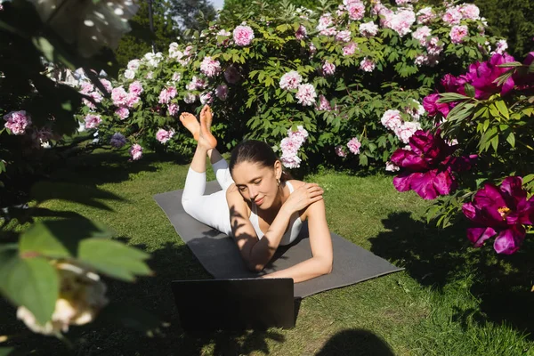 Sportliche Barfußfrau Liegt Auf Yogamatte Neben Laptop Und Blühendem Gebüsch — Stockfoto