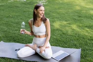 Mutlu kadın elinde şişeyle yoga minderinde oturuyor Lotus poz veriyor dizüstü bilgisayarın yanında 