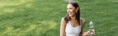 happy woman in white sports bra holding sports bottle and looking away outdoors, banner