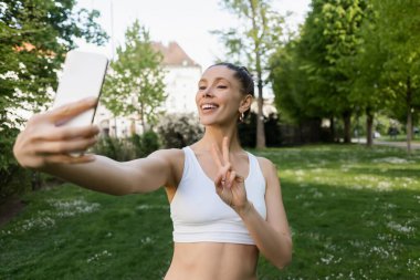 Beyaz spor sutyenli neşeli kadın zafer jesti yapıyor ve parkta bulanık akıllı telefondan selfie çekiyor.