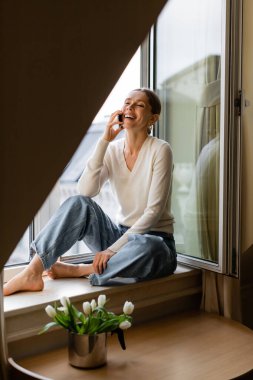 cheerful barefoot woman in jeans sitting on windowsill near tulips and talking on mobile phone