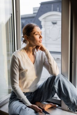 dreamy woman looking away while sitting on windowsill at home clipart