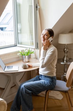 cheerful woman looking at camera while talking on cellphone near laptop and fresh tulips