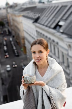 woman covered with shawl holding clay cup and smiling at camera near rooftops on blurred background clipart