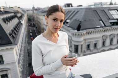 woman in white jumper holding clay cup and looking at camera on rooftop of city building