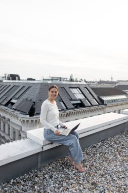 cheerful woman in jeans sitting on rooftop with laptop and smiling at camera
