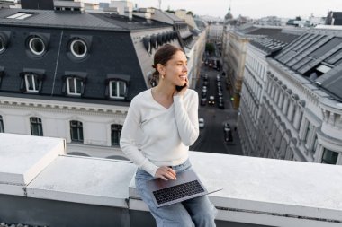 cheerful woman sitting on rooftop with laptop and talking on mobile phone