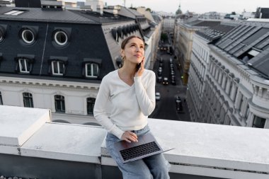 positive woman looking up while sitting on rooftop with computer and talking on cellphone