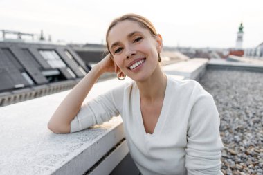 joyful woman smiling at camera while sitting on rooftop