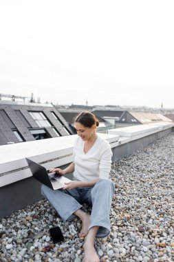smiling barefoot woman working on laptop while sitting on rooftop near mobile phone