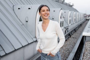 happy woman in white jumper standing with hands in pockets on rooftop