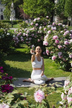 cheerful woman holding sports bottle while sitting in shoelace pose on lawn near blossoming peonies