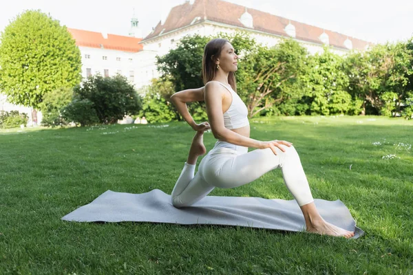 Beyaz taytlı bir kadının parkta eğri büğrü maymun pozunda yoga çalışmasının yan görüntüsü.