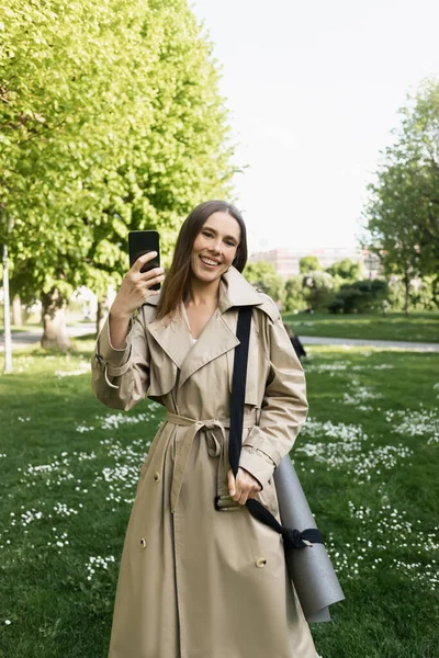 トレンチコートを着た幸せな女性がフィットネスマットで立ち 公園でスマートフォンで自撮り — ストック写真