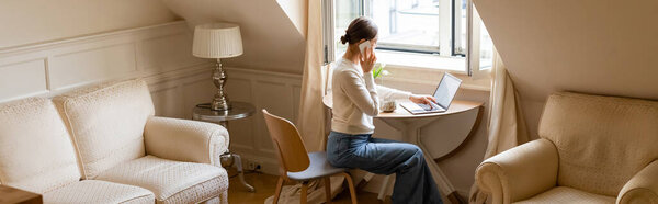 женщина в джинсах разговаривает на смартфоне возле ноутбука и окна в мансарде, баннер