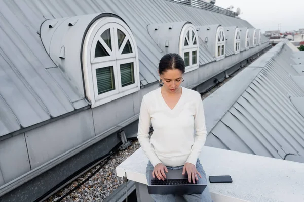 穿着白色套头毛衣的女人在智能手机旁边的笔记本电脑上打字 屋顶上的屏幕是空白的 — 图库照片