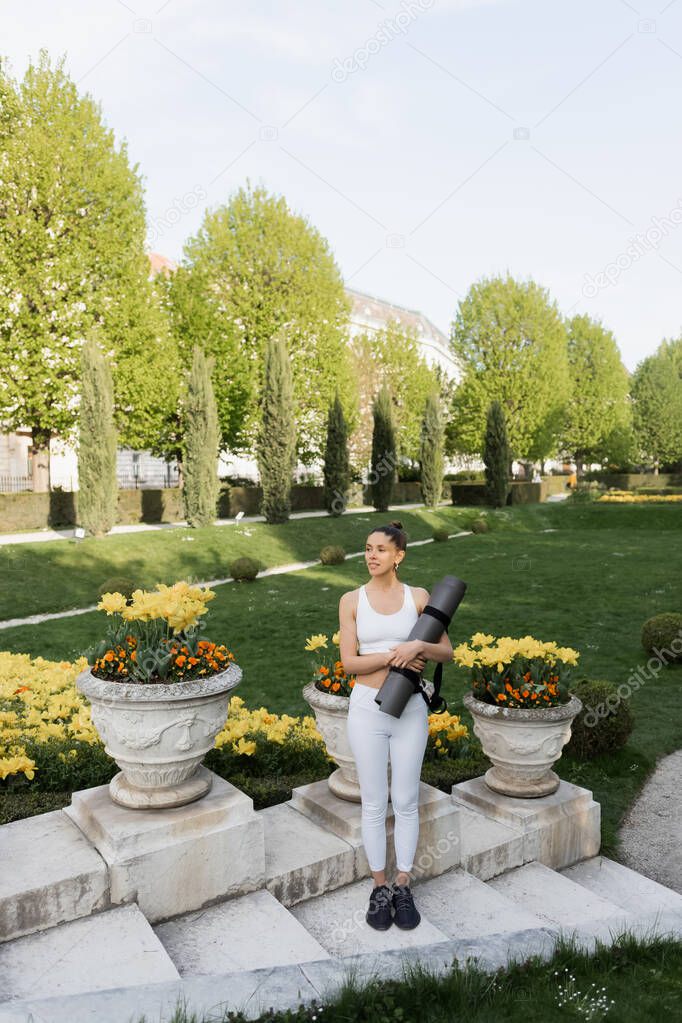 full length of woman in white sportswear standing with fitness mat near park vases with flowers