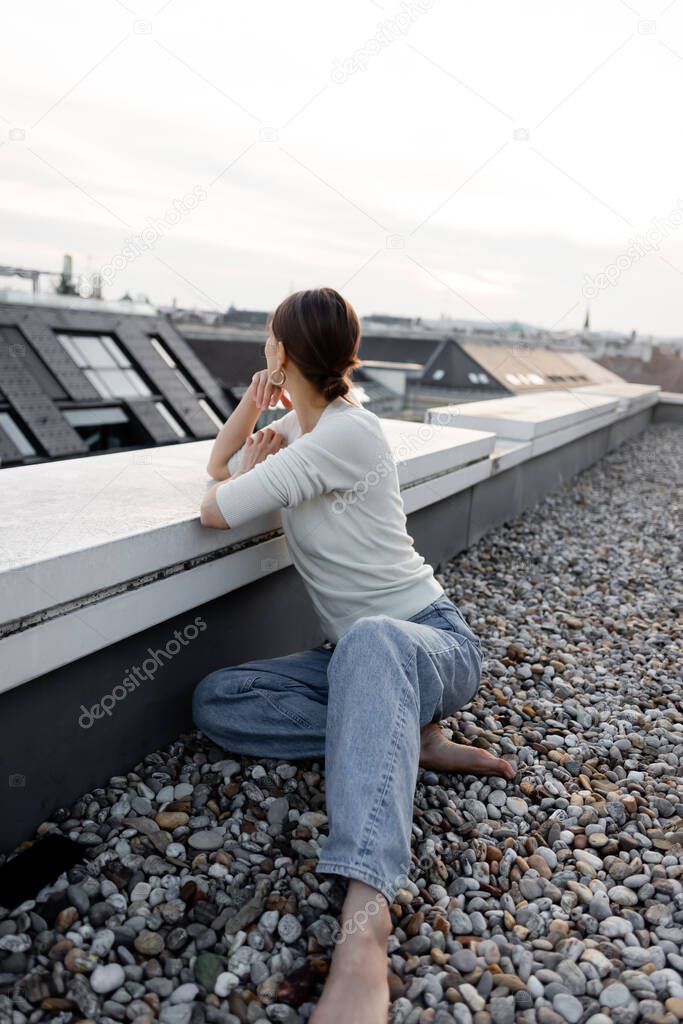 barefoot woman in jeans sitting on rooftop and looking away