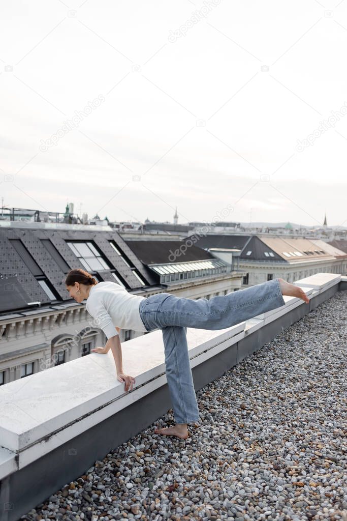 full length of barefoot woman in jeans looking down rooftop of urban building 