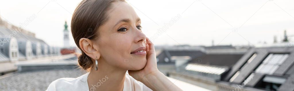 dreamy and smiling woman holding hand near face while looking away outdoors, banner