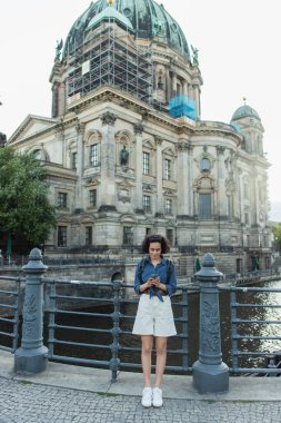 BERLIN, GERMANY - JULY 14, 2020: young curly tourist using smartphone in front of berlin cathedral 