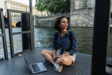 surprised freelancer holding smartphone while sitting with crossed legs near laptop and river in berlin 