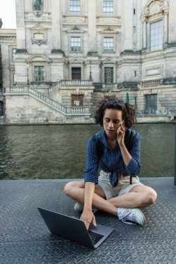 curly freelancer talking on smartphone while sitting with crossed legs near laptop and river in berlin 