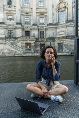 curly woman talking on smartphone while sitting with crossed legs near laptop and river in berlin 