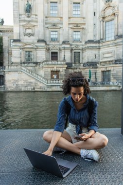 curly young woman using smartphone while sitting with crossed legs near laptop and river in berlin 