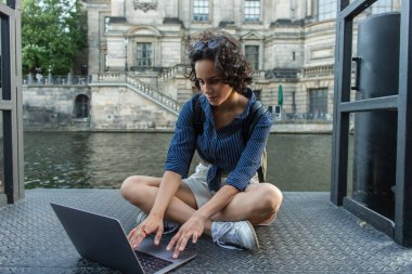 curly young woman using laptop while sitting with crossed legs near river in berlin 