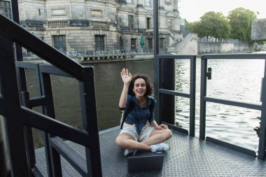 curly young woman having video call while sitting with crossed legs near river in berlin 