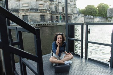 happy young woman using laptop while sitting with crossed legs near river in berlin 