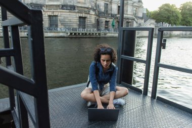 curly young woman in sunglasses using laptop while sitting with crossed legs near river in berlin 