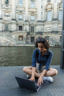 young freelancer using laptop while sitting with crossed legs near river in berlin 