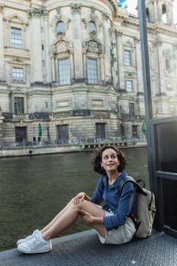 amazed and young woman sitting near river and ancient building in berlin 