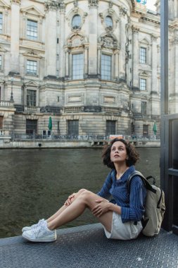 curly young woman sitting near river and ancient building in berlin 