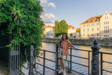 full length of curly woman in summer outfit standing near river in berlin 