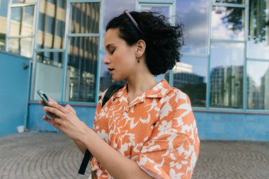 curly young woman messaging on smartphone near building in berlin  clipart