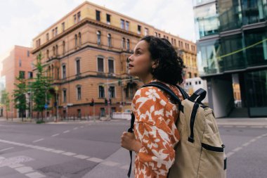 curly young woman with backpack standing on urban street in berlin 