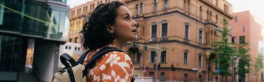 curly young woman with backpack standing on urban street in berlin, banner