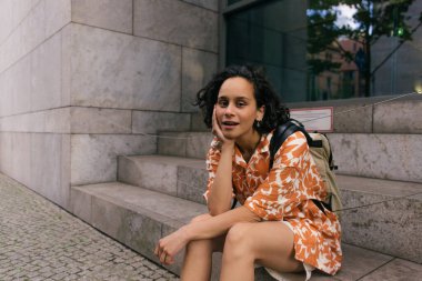 curly young woman in floral shirt sitting on stairs on street in berlin 