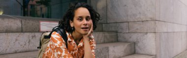 curly young woman in floral shirt sitting on stairs on street in berlin, banner