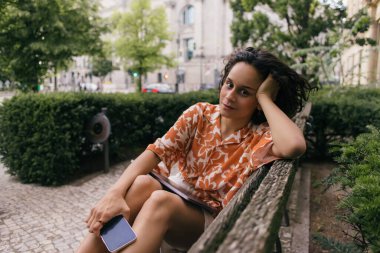 young woman in summer outfit holding smartphone and sitting on bench in green park 