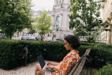 side view of young freelancer sitting on bench and using laptop in green park 